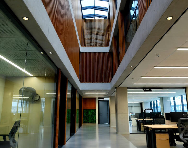 Et opdateret og stilfuldt interiør i et erhvervscenter med højt glasloft, naturligt lys, grønne planter og åbne etager med glasgelændere. Designet kombinerer beton, træ og glas med rene linjer, varm belysning og et mere kreativt præg, der skaber en inspirerende og professionel arbejdsatmosfære.